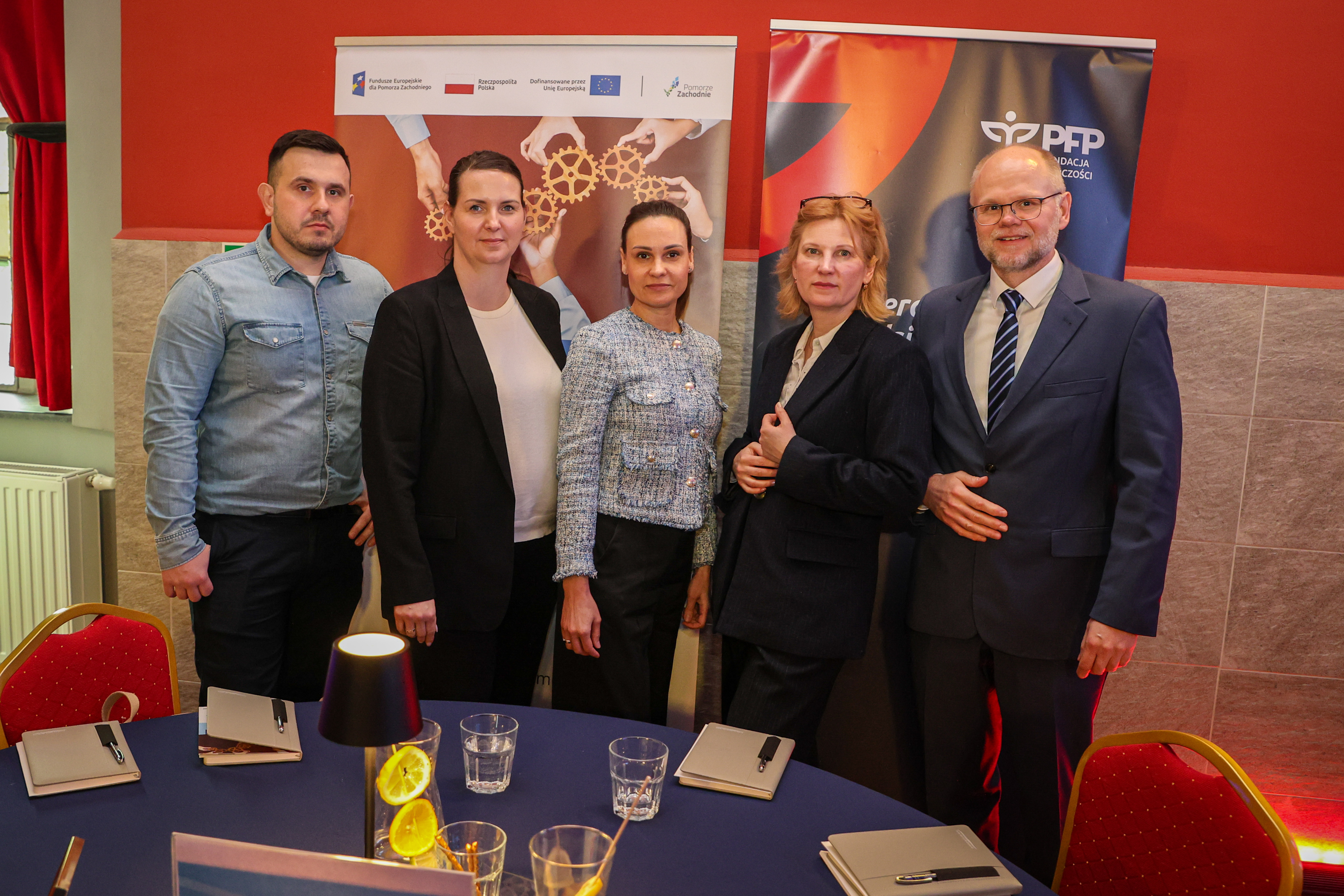 Pięć osób w strojach biznesowych stoi przy stole konferencyjnym na tle dwóch roll-upów i komunikuje spotkanie lub współpracę w obszarze funduszy i przedsiębiorczości, na lewym banerze napis Fundusze Europejskie dla Pomorza Zachodniego i grafika zębatek w dłoniach, na prawym logo PFP Polska Fundacja Przedsiębiorczości