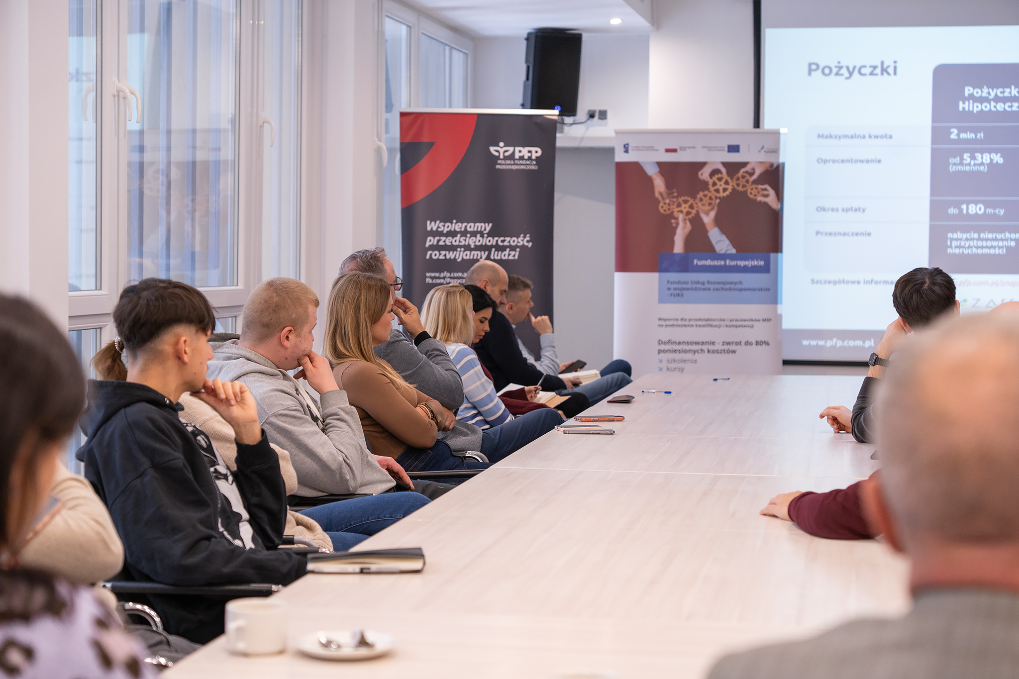 Na zdjęciu znajduje się grupa osób przy stole konferencyjnym. W tle widać roll upy.