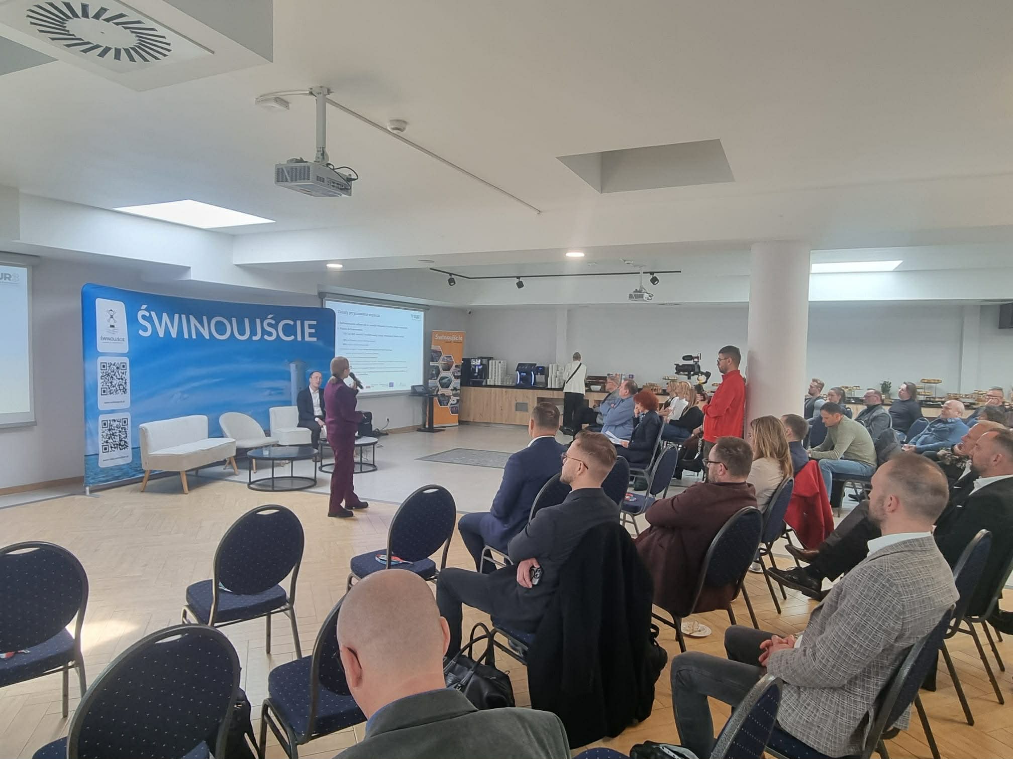 Sala konferencyjna z uczestnikami siedzącymi na krzesłach i prelegentem prowadzącym wystąpienie przy ekranie prezentacji, po lewej duży baner z napisem Świnoujście i kodami QR. Komunikat dotyczy spotkania informacyjnego lub konferencji dla przedsiębiorców.