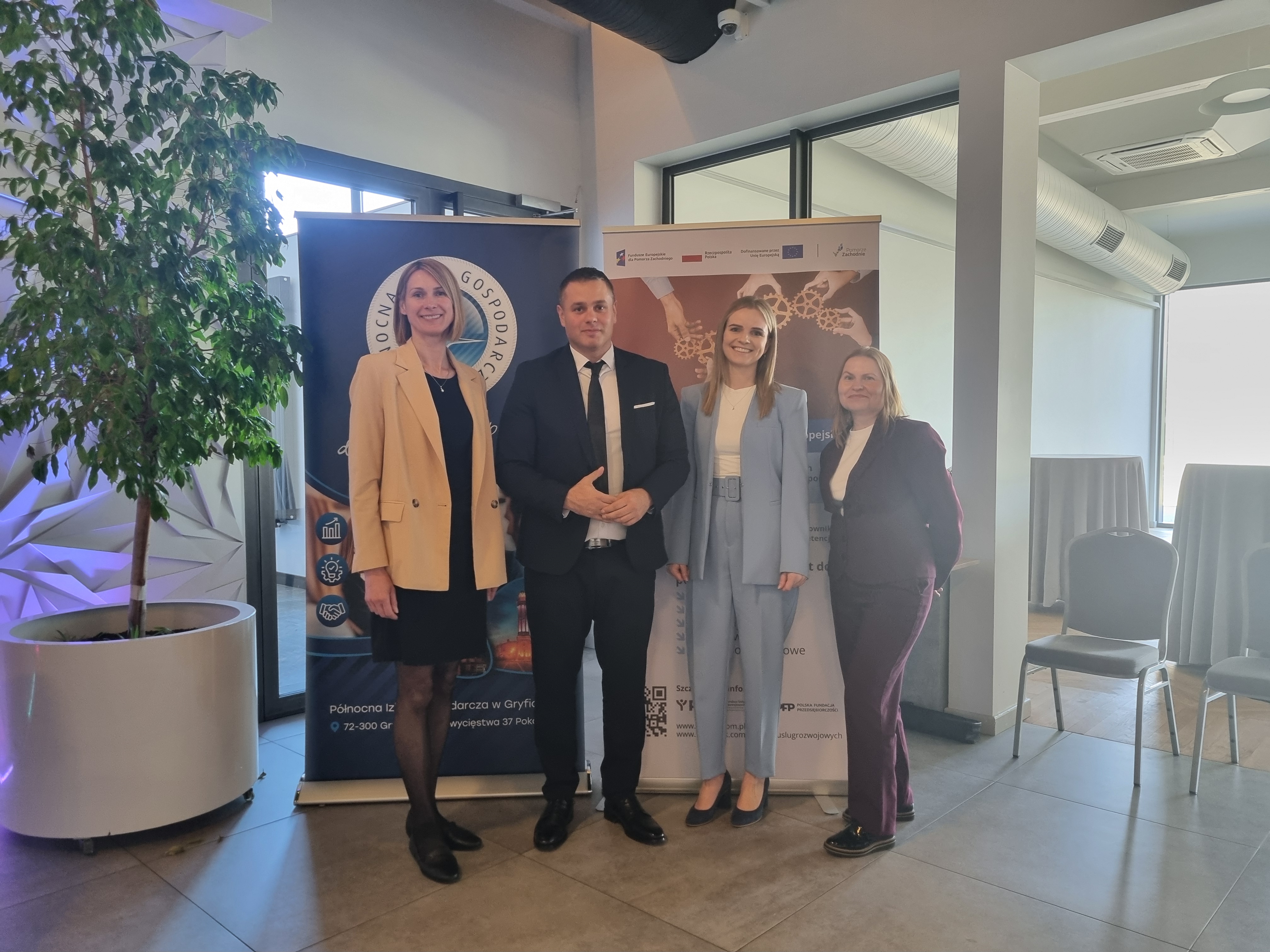 Cztery osoby stoją w sali konferencyjnej przed roll-upami promującymi instytucje i programy wsparcia, komunikat o współpracy i działaniach na rzecz przedsiębiorczości